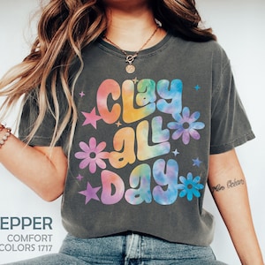 May include: A dark grey t-shirt with a tie-dye effect graphic that says "Clay All Day" in rainbow colors. The graphic is surrounded by stars and flowers.
