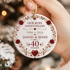 Puede incluir: Un adorno redondo de cerámica blanca con una cinta dorada. El diseño incluye rosas rojas, filigrana dorada y un corazón rojo central. El texto dice: OUR RUBY ANNIVERSARY, 1986-2026, DANIEL & RENEE y 40 YEARS en letras de color rojo oscuro.