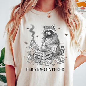 Puede incluir: Camiseta color marfil con un diseño de mapache alfarero y el texto "FERAL & CENTERED". El mapache lleva un delantal y trabaja en un torno de alfarero. El diseño incluye estrellas, un horno y herramientas de alfarería.