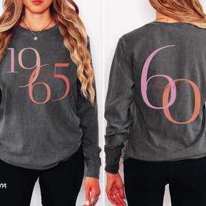 May include: A long-sleeved shirt in a dark grey color with a pink and orange design. The front of the shirt has the number "1965" with the words "next chapter" written in a script font. The back of the shirt has the number "60" with the words "next chapter" written in a script font.