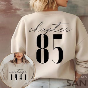 Op de afbeelding: Een zandkleurige sweatshirt met de tekst "chapter 85" in zwart script en cijfers op de rug. Een kleinere afbeelding op de voorkant toont een vrouw die een vergelijkbare sweatshirt draagt met de tekst "vintage 1941".