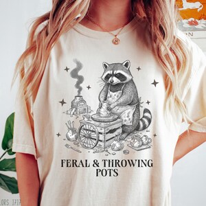 Può includere: T-shirt color avorio con un disegno di procione vasaio e la scritta "FERAL & THROWING POTS". Il procione è raffigurato a un tornio, con un grembiule, attrezzi da vasaio e un forno. La maglietta è una Comfort Colors 1717.