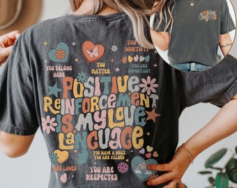 May include: A dark grey t-shirt with a colorful graphic design that says "Positive Reinforcement is my love language". The graphic includes hearts, stars, flowers, and rainbows. The text on the shirt also includes phrases like "You belong here", "You matter", "You are important", "You are loved", "You are respected", and "You have a voice, you are valued".