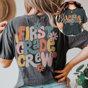 May include: A black t-shirt with a retro-style graphic that says "First Grade Crew" in colorful letters. The graphic also includes a pencil, an apple, and flowers.