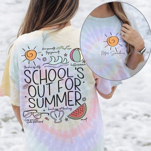 May include: A tie-dye t-shirt with a rainbow colour scheme. The shirt has a graphic design with the text "School's Out For Summer" and various summer-themed illustrations, including a sun, a beach ball, a watermelon, and a seashell. The text "Mrs. Sanders" is also visible on the shirt.