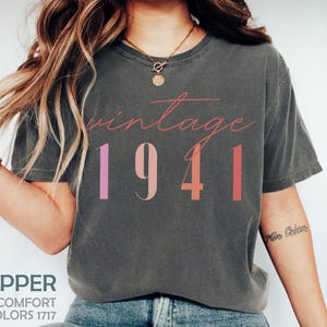 Puede incluir: Camiseta gris oscuro con la palabra "vintage" en cursiva sobre los números "1941". Los números son en tonos rosa y coral. La camiseta también tiene las palabras "Pepper", "Comfort Colors 1717" impresas en la parte inferior.