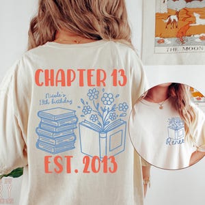 May include: Ivory-colored t-shirt with a graphic design. The design includes the text "CHAPTER 13" in coral, along with "EST. 2013". A stack of books and an open book with flowers are also featured in blue. The shirt is a Comfort Colors brand.