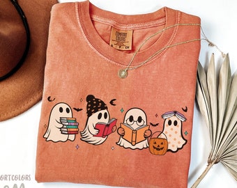 May include: A peach-colored t-shirt with a graphic of four ghosts reading books. The ghosts are wearing hats and glasses. One ghost is holding a pumpkin. The text "COMFORT COLORS" is on the tag.