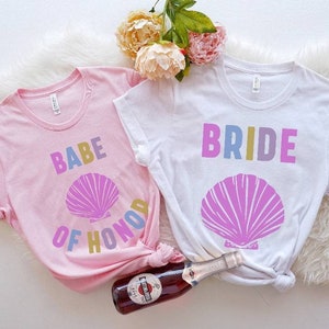 May include: Two white t-shirts with a pink seashell graphic. One shirt says "BRIDE" in rainbow colors and the other says "BABE OF HONOR" in rainbow colors.