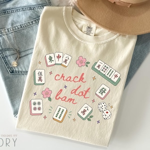 May include: An ivory-coloured t-shirt featuring a graphic print of various mahjong tiles, pink cherry blossoms, and small stars. The text "crack dot bam" is printed in a cursive red font in the centre. The shirt has a crew neckline and short sleeves.