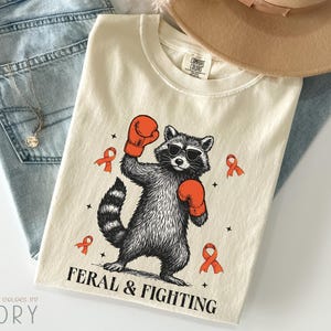 May include: Ivory-colored t-shirt featuring a raccoon wearing sunglasses and boxing gloves, with the text "FERAL & FIGHTING". The design includes orange ribbons and stars. The shirt is laid on top of blue jeans and a tan hat.