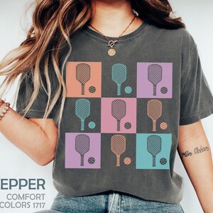 May include: Dark grey t-shirt with a grid design of pickleball paddles and balls in pastel shades. The shirt includes the text "PEPPER" and "COMFORT COLORS 1717". A relaxed, athletic style.