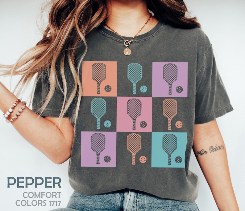 May include: Dark grey t-shirt with a grid design of pickleball paddles and balls in pastel shades. The shirt includes the text "PEPPER" and "COMFORT COLORS 1717". A relaxed, athletic style.