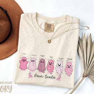 May include: A cream-colored t-shirt featuring a graphic of six pink ghosts arranged in a row, each representing a different level of pain. The text reads "Pain Scale" with labels above each ghost. The shirt has a Comfort Colors brand neck label.