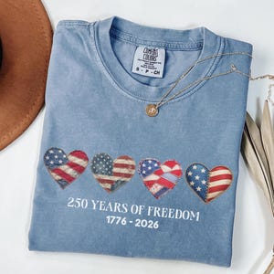 Op de afbeelding: Een lichtblauw T-shirt met vier hartvormige patches met het ontwerp van de Amerikaanse vlag. De tekst "250 YEARS OF FREEDOM 1776-2026" is eronder gedrukt. Een gouden ketting ligt over het shirt.