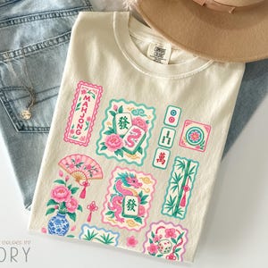 May include: An ivory t-shirt featuring a colourful graphic print of mahjong tiles, a pink dragon, a folding fan, a blue and white vase with pink flowers, bamboo stalks, and dice. The shirt has a crew neckline and a Comfort Colors brand label.
