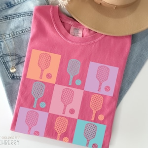 May include: A pink Comfort Colors t-shirt featuring a grid design of pickleball paddles and balls in different colours. The shirt is folded on top of blue jeans and a tan hat.
