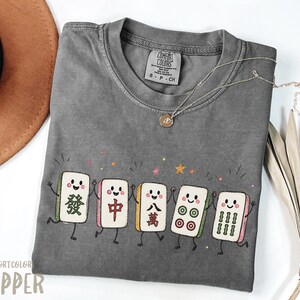 May include: A charcoal grey short-sleeve t-shirt featuring a graphic of five smiling Mahjong tiles holding hands. The tiles display various traditional symbols and colours. The shirt has a crew neckline and a Comfort Colors brand label inside.