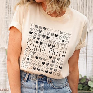 Pode incluir: Camiseta creme claro com um quadrado de cora&ccedil;&otilde;es pretos e brancos e as palavras "SCHOOL PSYCH" em preto. A camiseta &eacute; combinada com jeans azuis.