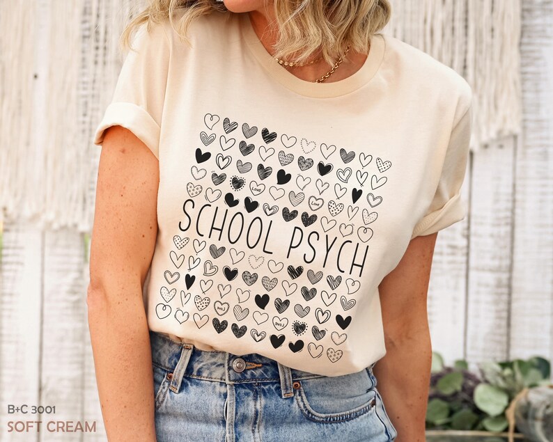 Pode incluir: Camiseta creme claro com um quadrado de cora&ccedil;&otilde;es pretos e brancos e as palavras "SCHOOL PSYCH" em preto. A camiseta &eacute; combinada com jeans azuis.