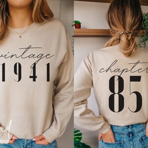 Könnte beinhalten: Sandfarbener Pullover mit dem Aufdruck "vintage 1941" in schwarzer Schrift und Blockbuchstaben auf der Vorderseite. Auf der Rückseite steht "chapter 85" in schwarzer Schrift.