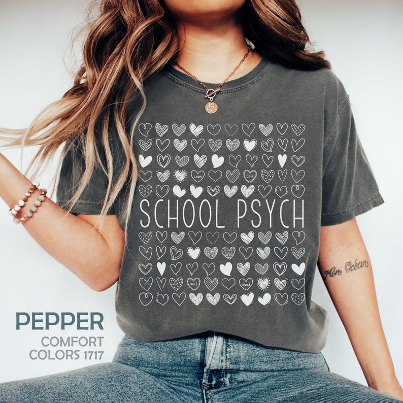 Pode incluir: Uma t-shirt cinza escuro com um desenho de cora&ccedil;&atilde;o branco e as palavras "SCHOOL PSYCH". O desenho &eacute; composto por muitos cora&ccedil;&otilde;es pequenos desenhados &agrave; m&atilde;o. A t-shirt tamb&eacute;m tem as palavras "PEPPER" e "COMFORT COLORS 1717".