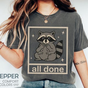 May include: Pepper-colored t-shirt featuring a raccoon illustration within a square frame, with the words "all done" below. The raccoon is gray and white, with a striped tail. The shirt also has the words "Comfort Colors 1717".