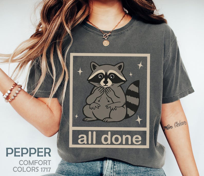 May include: Pepper-colored t-shirt featuring a raccoon illustration within a square frame, with the words "all done" below. The raccoon is gray and white, with a striped tail. The shirt also has the words "Comfort Colors 1717".