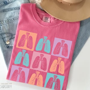 May include: A pink t-shirt with a crew neck featuring a pattern of colorful lungs in squares. The lungs are in shades of orange, teal, and pink. The shirt is folded on top of a pair of blue jeans and a tan hat.