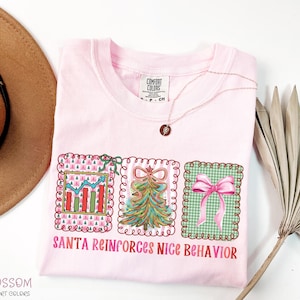 Op de afbeelding: Een lichtroze Comfort Colors T-shirt met de tekst "SANTA REINFORCES NICE BEHAVIOR". De afbeelding bevat een grafiek, een kerstboom en een roze strik. Een gouden ketting ligt over het shirt. Het shirt ligt op een witte ondergrond.