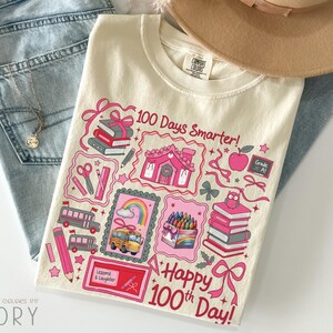 May include: Ivory-colored t-shirt with a colorful graphic celebrating the 100th day of school. The design includes pink illustrations of books, a school bus, a house, and the text "100 Days Smarter!" and "Happy 100th Day!"
