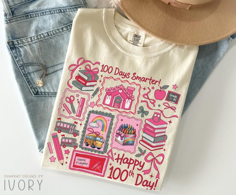 May include: Ivory-colored t-shirt with a colorful graphic celebrating the 100th day of school. The design includes pink illustrations of books, a school bus, a house, and the text "100 Days Smarter!" and "Happy 100th Day!"