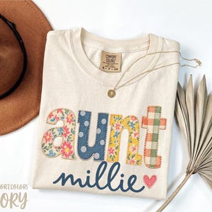 Puede incluir: Una camiseta de color marfil con la palabra aunt en letras de tela estampada de colores con borde cosido. Debajo, el nombre millie está impreso en letra cursiva azul oscuro junto a un pequeño corazón rojo. La camiseta tiene una etiqueta Comfort Colors.