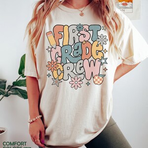 May include: A white t-shirt with the text "First Grade Crew" in colorful retro lettering. The text is surrounded by stars, flowers, and a pencil.