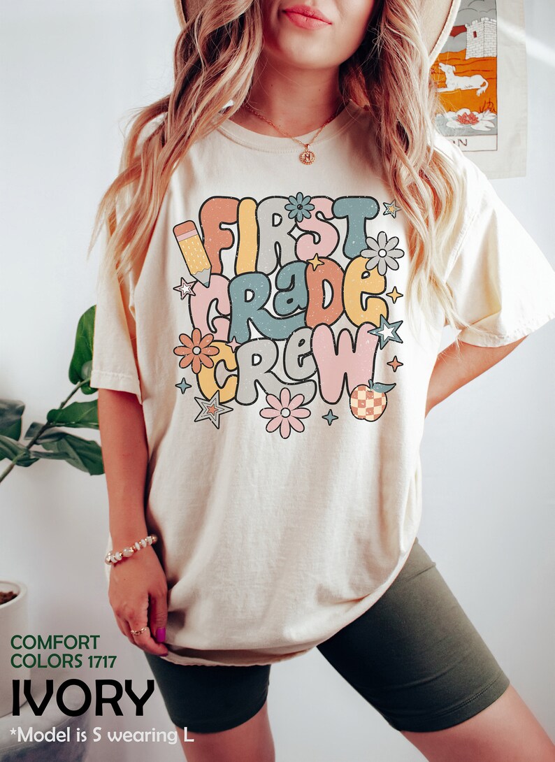 May include: A white t-shirt with the text "First Grade Crew" in colorful retro lettering. The text is surrounded by stars, flowers, and a pencil.