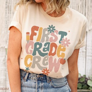 May include: A cream colored t-shirt with the text "First Grade Crew" in colorful, retro-style lettering. The text is surrounded by small stars, flowers, and a pencil and apple graphic.