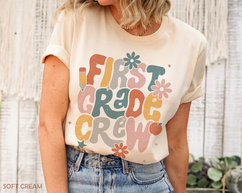 May include: A cream colored t-shirt with the text "First Grade Crew" in colorful, retro-style lettering. The text is surrounded by small stars, flowers, and a pencil and apple graphic.