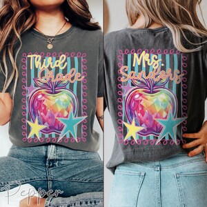 May include: A charcoal gray t-shirt featuring a graphic print of a colorful, faceted apple surrounded by a pink border, teal stripes, and yellow and teal stars. The front displays the text Third Grade, while the back displays Mrs. Sanders.