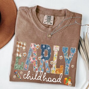 May include: Brown t-shirt featuring the words "EARLY childhood" in a patchwork design. The letters are made of various floral and patterned fabrics. A pencil and a gift are also featured. A silver necklace with a pendant is on the shirt.