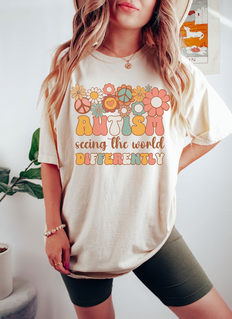 May include: A cream colored t-shirt with a retro-style graphic featuring flowers, peace signs, and the text "AUTISM seeing the world DIFFERENTLY".
