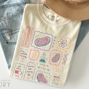 Könnte beinhalten: Ein elfenbeinfarbenes T-Shirt mit einem bunten, wissenschaftlich gestalteten Grafikdruck. Das Design zeigt Illustrationen von Mitochondrien, einem Atom, einem Becherglas, einem DNA-Strang und einem Blitz sowie Text wie Mitochondria, ATP loading und chemische Formeln.