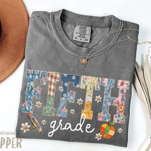 Puede incluir: Una camiseta gris Comfort Colors con las palabras "FIFTH grade" en un diseño de patchwork. Las letras están hechas de varias telas florales y estampadas. Un lápiz y una calabaza también aparecen en el diseño.