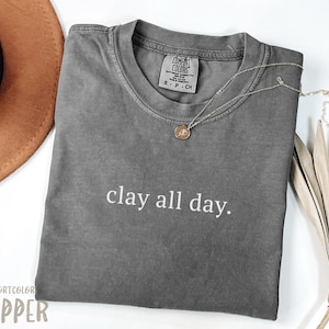 Puede incluir: Camiseta gris con el texto "clay all day." impreso en blanco en la parte delantera. La camiseta está hecha de un material suave y cómodo.