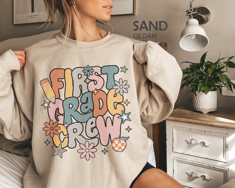 May include: A sand-colored sweatshirt with a retro-style graphic that says "First Grade Crew" in colorful letters. The graphic also includes a pencil, stars, and flowers.