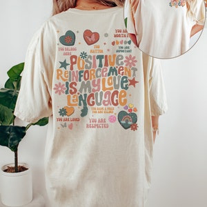 Pode incluir: Camiseta de cor marfim com uma estampa gr&aacute;fica colorida nas costas que diz "Positive Reinforcement is my love language". O gr&aacute;fico inclui cora&ccedil;&otilde;es, estrelas, borboletas e arco-&iacute;ris. A frente da camiseta tem um pequeno gr&aacute;fico que diz "Mrs. Sanders".