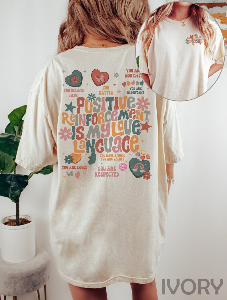 Pode incluir: Camiseta de cor marfim com uma estampa gr&aacute;fica colorida nas costas que diz "Positive Reinforcement is my love language". O gr&aacute;fico inclui cora&ccedil;&otilde;es, estrelas, borboletas e arco-&iacute;ris. A frente da camiseta tem um pequeno gr&aacute;fico que diz "Mrs. Sanders".