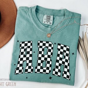 Op de afbeelding: Een lichtgroen T-shirt met een zwart-wit geruite "ABA"-grafiek. Het shirt is in vintage stijl, met een verweerde look. Het shirt is gemaakt door Comfort Colors.