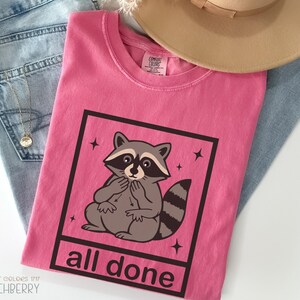 May include: A pink t-shirt with a raccoon graphic inside a black square, with the words "all done" below. The shirt is folded on top of blue jeans and a gold necklace. A tan hat is also visible.