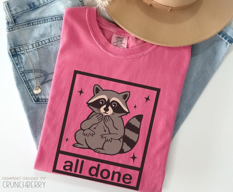 May include: A pink t-shirt with a raccoon graphic inside a black square, with the words "all done" below. The shirt is folded on top of blue jeans and a gold necklace. A tan hat is also visible.