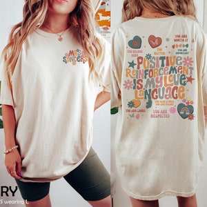 Pode incluir: Camiseta oversized de cor marfim com um design floral nas costas que diz "Refor&ccedil;o positivo &eacute; a minha linguagem de amor". A frente da camiseta tem um pequeno design floral com o texto "Mrs. Sanders".